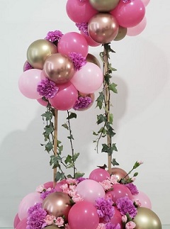Cluster of Pink and gold balloons with pink flowers stuffed in between the balloons on 2 sticks and tied to a basket with pink and gold balloons similar as on top